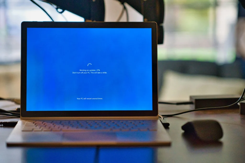 A laptop displays a Windows update screen showing the system installing updates at 27%, illustrating the process of updating software and hardware drivers on an older computer.