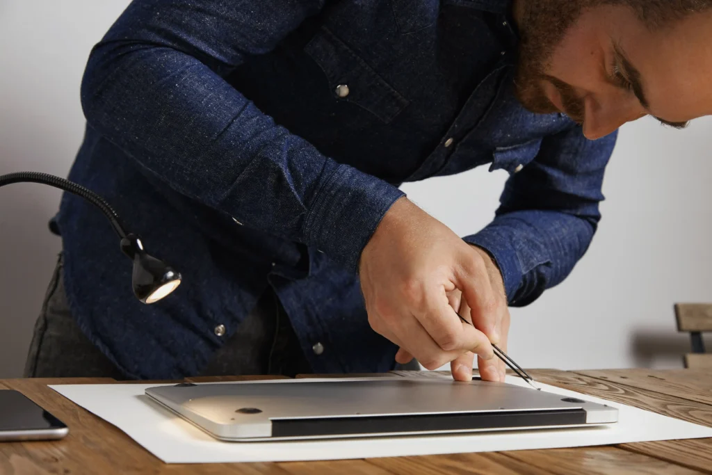 Technician using tweezers to secure a small screw on the underside of a laptop, finishing the repair process on a wooden workbench.