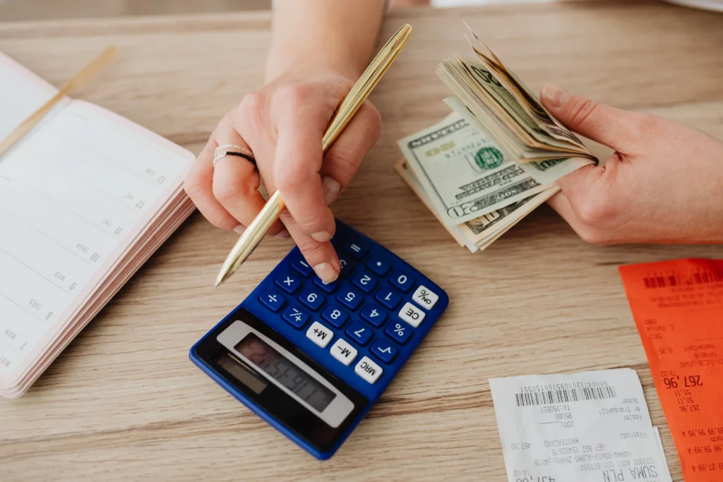 Person calculating laptop repair expenses with a calculator while reviewing bills, receipts, and cash to compare repair costs with the cost of replacing a laptop.