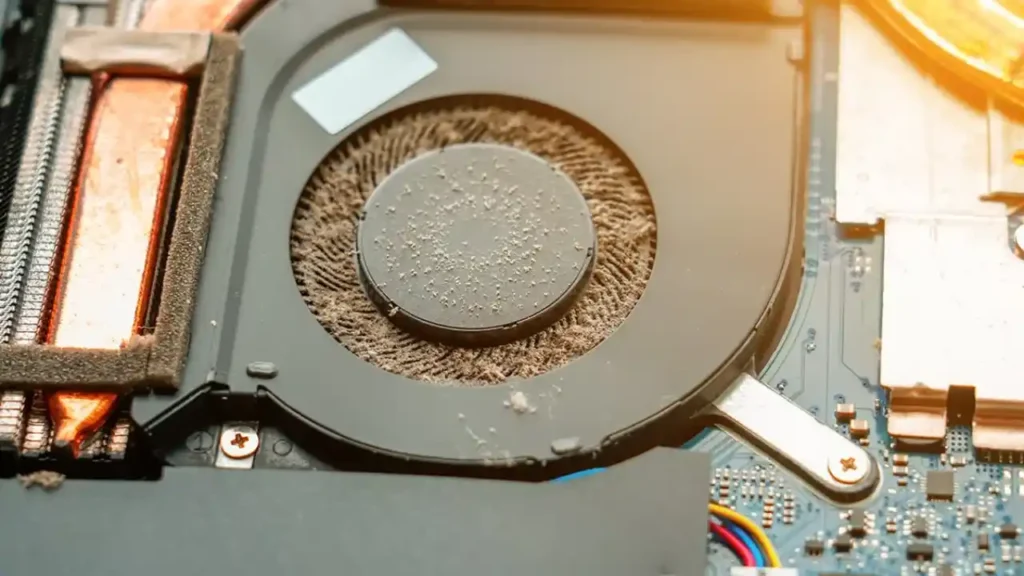 Close-up of a dusty computer fan inside a laptop, surrounded by copper and aluminum components. The image conveys neglect and the need for cleaning.