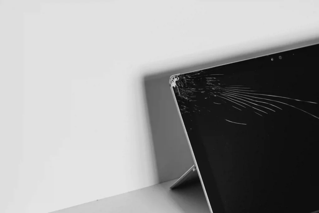 Cracked laptop display screen requiring hardware repair leaning against a white wall.