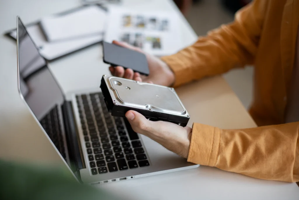 A person in a mustard-yellow shirt holds a standard internal hard disk drive and a portable external storage drive beside an open laptop on a white desk. The image illustrates different storage devices used for transferring files, backing up data and expanding computer storage.