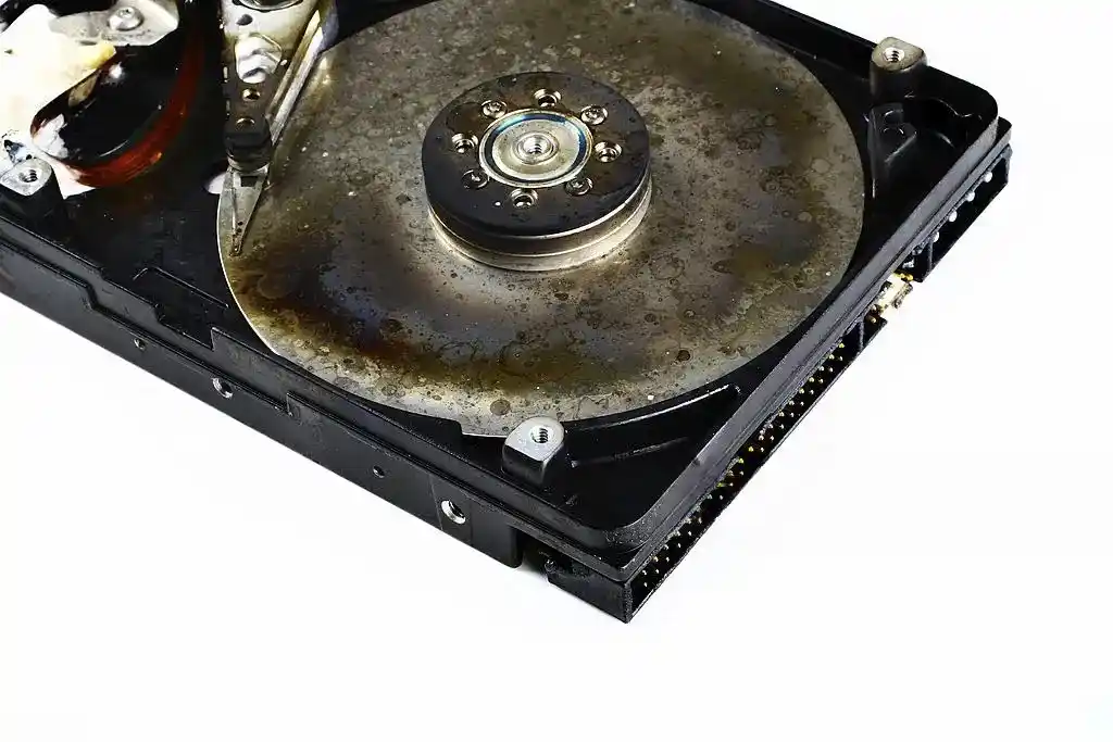 Close-up of an opened hard disk drive showing a visibly discoloured and stained metal platter, central spindle and internal mechanical components. The black drive casing and connector pins are visible against a white background. The image illustrates physical hard drive damage, platter deterioration and potential storage device failure.
