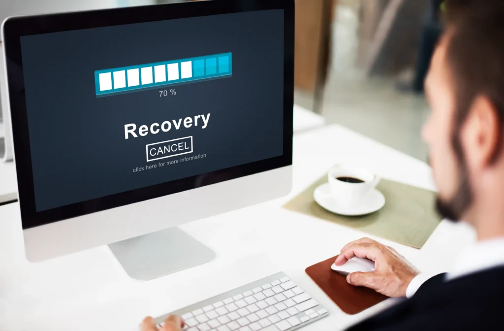 A person monitoring a data recovery process on a desktop computer, with the screen displaying a recovery progress bar at 70%. The computer setup includes a keyboard, mouse and coffee cup on a clean white desk. The image represents data recovery, file restoration and the process of recovering lost or inaccessible data.