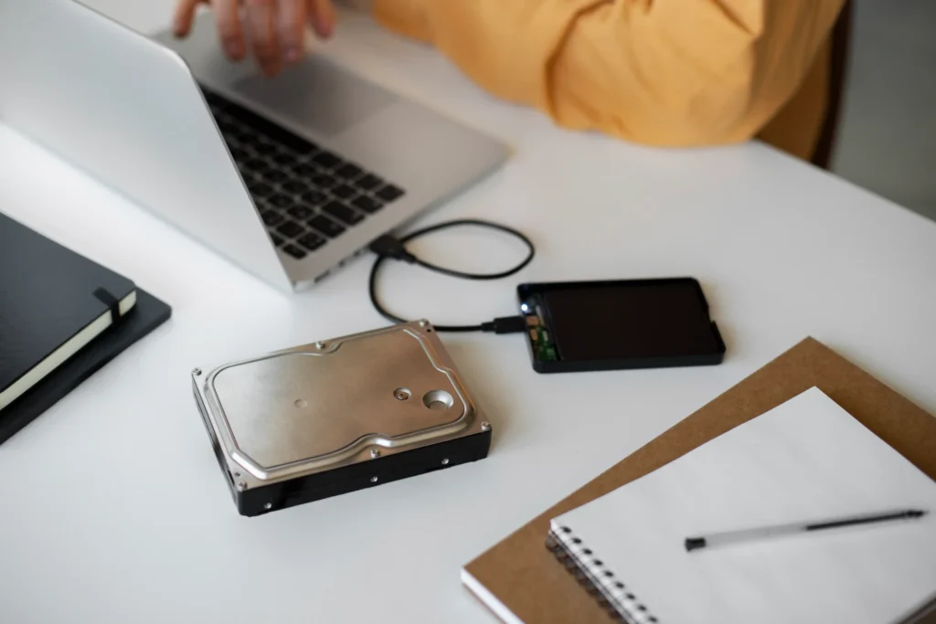 An external hard drive and a portable storage drive connected to a laptop on a white desk, illustrating external storage devices used for transferring, backing up and accessing digital files. A notebook, pen and black journal are also visible on the desk.