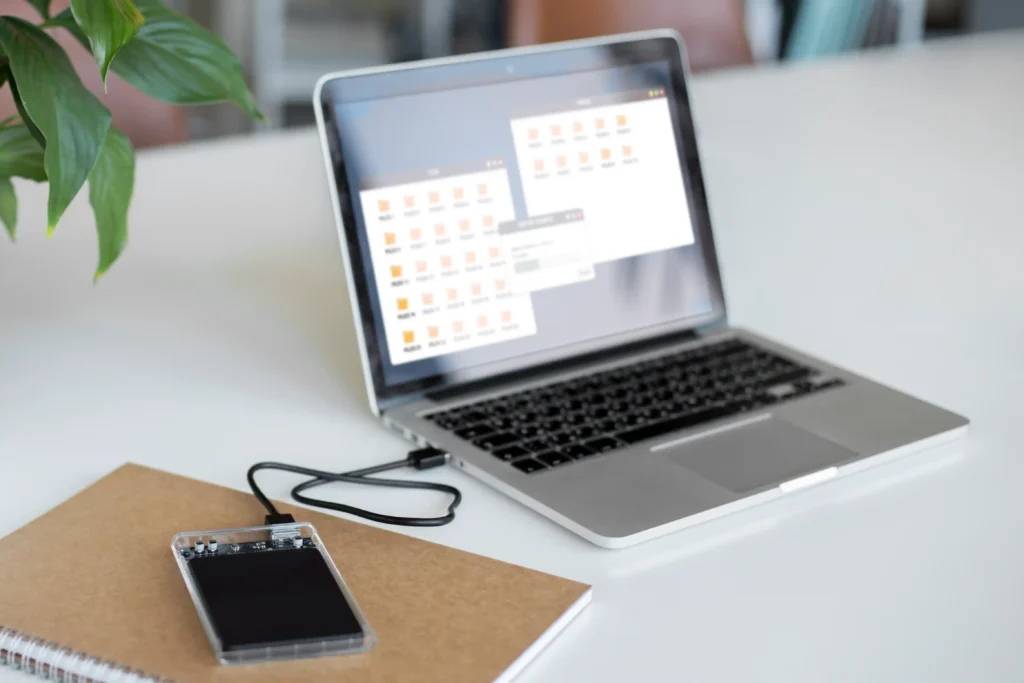 An external storage drive in a transparent enclosure connected to a silver laptop via a USB cable on a white desk. The laptop displays several file folders, illustrating how an external hard drive can be used to access, transfer and back up computer files. A notebook and green potted plant are visible nearby.