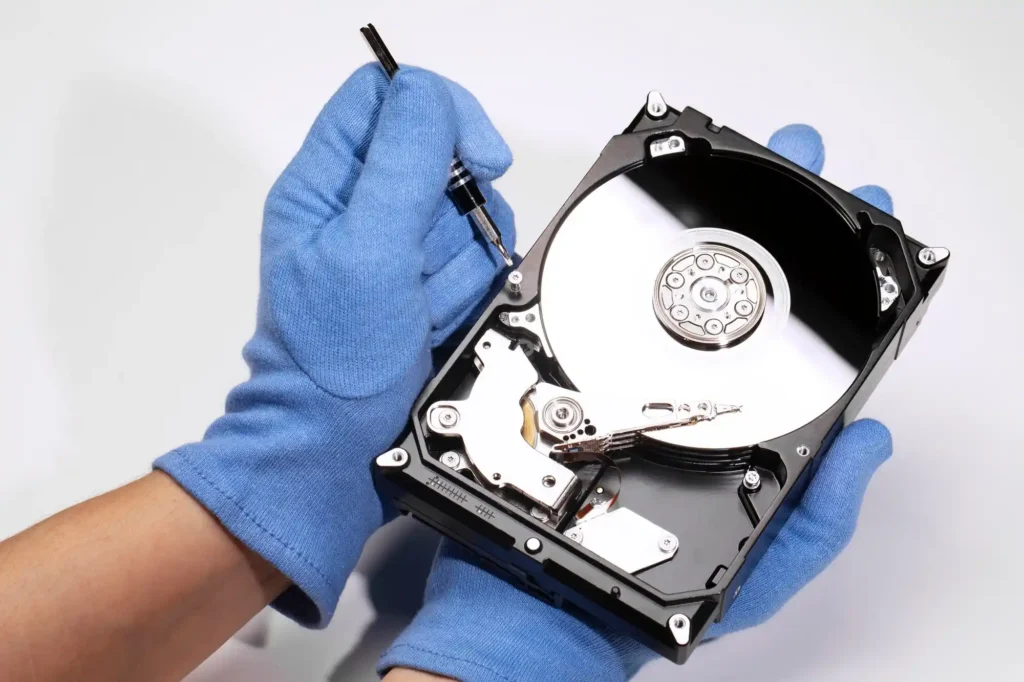 A technician wearing blue protective gloves carefully repairing an opened hard disk drive with a precision screwdriver. The exposed drive platter, actuator arm and internal components are visible against a clean white background. The image represents professional hard drive repair, internal component inspection and specialist data recovery procedures.