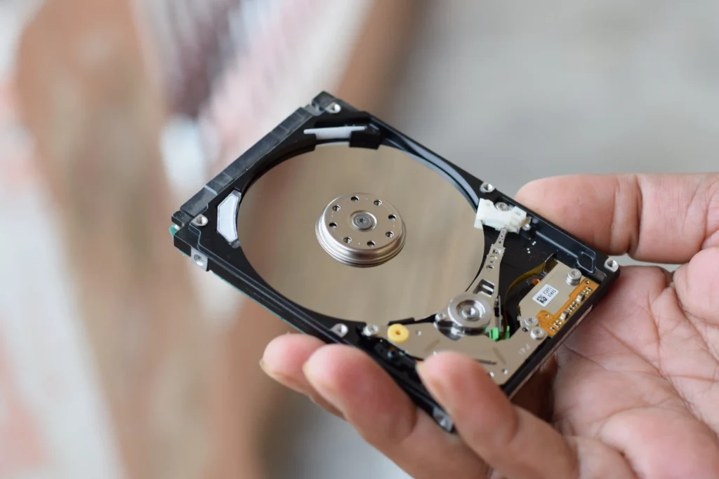 Close-up of a hand holding an opened hard disk drive (HDD), revealing the circular magnetic platter, central spindle, read/write actuator arm and internal electronic components. The exposed drive mechanism illustrates the internal structure of a hard drive used for data storage and recovery.