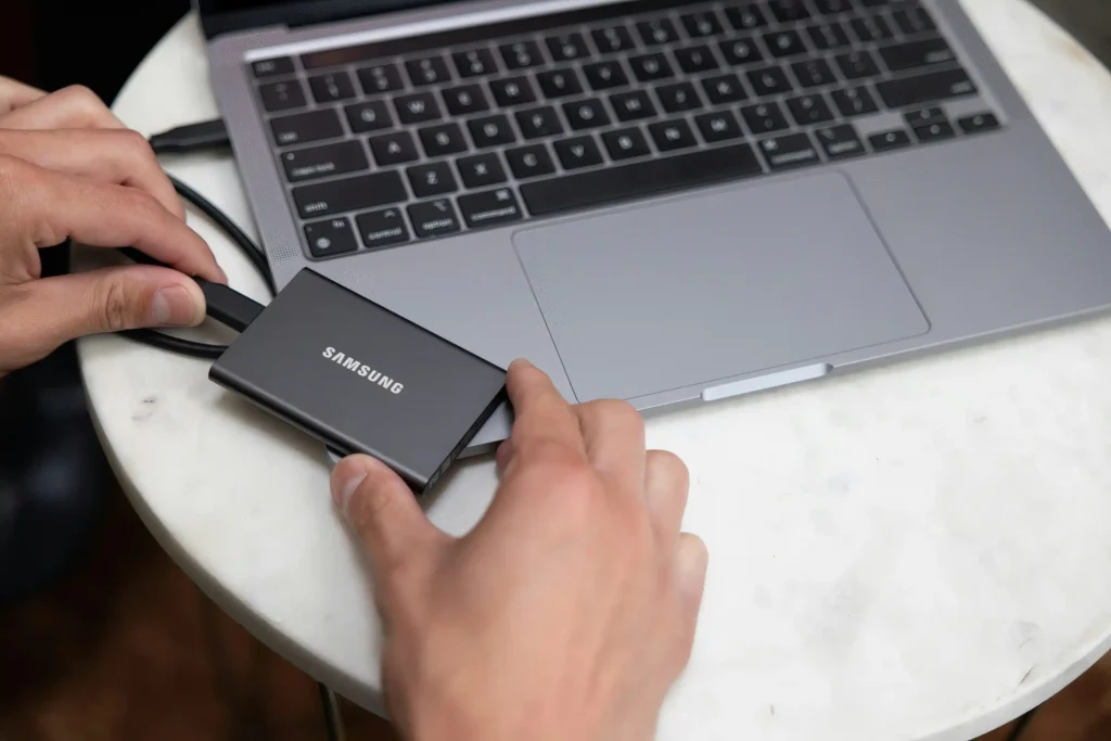 A person connects a Samsung portable solid-state drive (SSD) to a silver laptop using a USB cable on a white table. The close-up shows the external storage device being held beside the laptop's keyboard and trackpad, illustrating external SSD connectivity, file transfers and data backup.