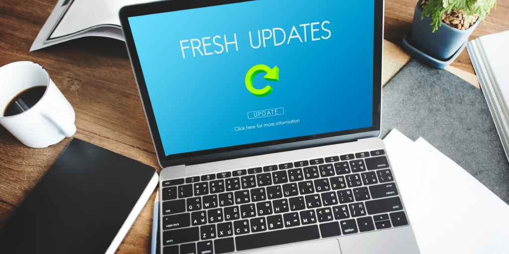A laptop displaying a software update screen with a blue background, green refresh icon and update button, viewed from above on a wooden desk. A coffee cup, notebook, papers and small potted plant surround the laptop. The image represents software updates, installing the latest version, and keeping applications up to date.
